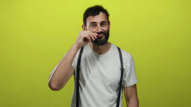 Sakallı İspanyol bir adam, beyaz gömlek ve pantolon askısı giymiş, özel hayat kavramı taşıyan parlak sarı arka plana karşı gizli bir jest yapıyor..