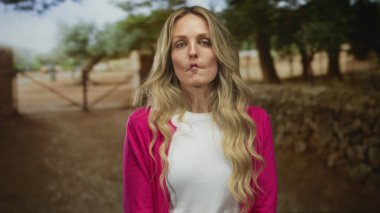 Parkta, güneşli bir günde yeşil ağaçlarla çevrili pembe kazak ve beyaz gömlek giyen bir kadın neşeli ve hayat dolu bir atmosfer getiriyor..