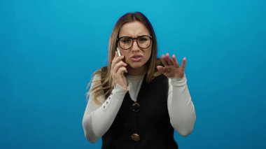 Telefonda kafası karışmış bir şekilde konuşan bir kadın, mavi bir arkaplan karşısında, gözlük takan ve serbest elle hareket eden, sürpriz ve konuşma yoğunluğunu ileten bir kadın..