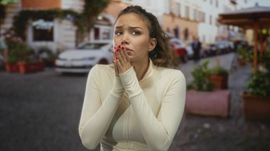 Genç İspanyol kadın göğsüne yakın ellerini çırpıyor ve park halindeki arabalar ve saksı bitkileriyle dolu caddeye bakıyor..