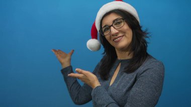 Gözlüklü gülümseyen İspanyol kadın stüdyoda elleri açık kırmızı Noel Baba şapkasını sunar; bayram neşesi.