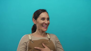 Parlak turkuaz bir duvara karşı bronz bir önlükle gülümseyen kadın neşeli bir ifadeyle dost bir atmosfer izlenimi veriyor..