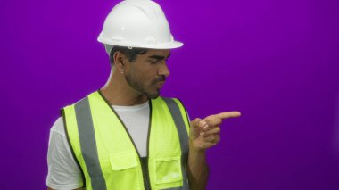 Beyaz kasketli ve floresan yelek giyen mühendis canlı mor stüdyo duvarına parmak işareti yapıyor..
