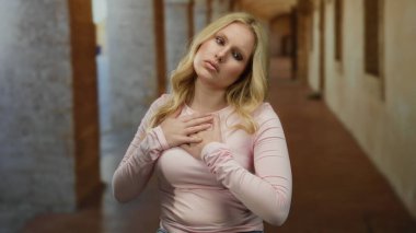 Üniversite koridorunda elleri göğsünde duygularını ifade eden bir kadın. Eski bir kampüs arka planında sarı saçlar ve düşünceli bir ifade sergiliyor..