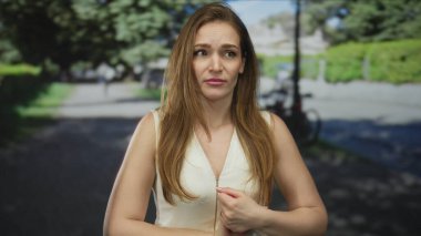 Parkta elleri sıkan ve beyaz kolsuz bluzuyla kaşlarını çatan kadın; endişe gerilimi korkusu.