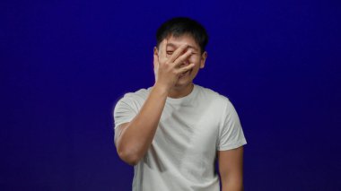 Mavi arka planda gülümseyen genç adam, eğlence ya da utanç ifade ediyor, günlük beyaz gömlek giyiyor..