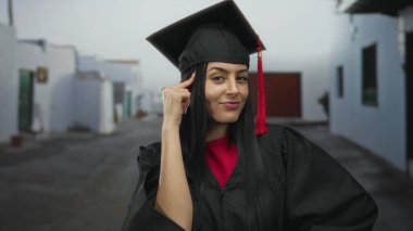 Mezuniyet şapkalı genç İspanyol kadın köyün arka planında ileri görüşlü ileri görüşlü akademik başarı ve gelecekteki olasılıklara olan güvenini gösteriyor.