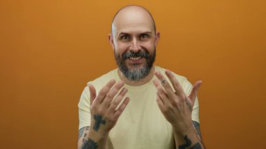 Kel sakallı adam, elleri turuncu duvara karşı hareketler yapıyor dövmeler ve davet edici ifadeler sergiliyor, izole bir stüdyoda sarı bir gömlek giyiyor..