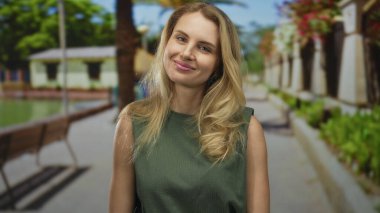 Yeşil ağaçlar ve yürüyüş yolu olan parkta gülümseyen bir kadın, güneşli bir günün tadını çıkaran genç sarışın, sakin ve sakin bir arka planda..