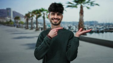 Gülümseyen süveter içindeki genç adam, açık gökyüzünün altında palmiye ağaçları ve marina arkaplanı olan deniz kenarındaki bir geçidi işaret ederek modern gündelik yaşam tarzını resmediyor..