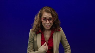 Gözlüklü, kırmızı bluzlu, bej ceketli, mavi arka planda duran, stüdyo ışıkları altında çeşitli ifadeler sergileyen bir kadın..