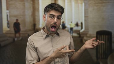 Gözlüklü genç İspanyol adam sıcak akşam ışığı altında tarihi bina cephesini işaret ediyor..