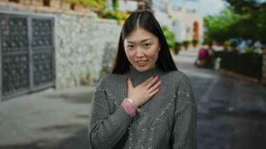 Şehir caddesinde açık havada, göğsünde gri bir kazak olan bir kadın şehir ortamında şaşkınlığını ifade ediyor.