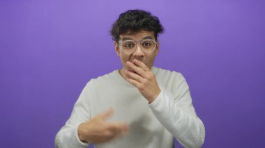 İspanyol adam mor stüdyo ortamında elleri ile ağız kaplar kameraya bakarken gözlük ve beyaz gömlek giyer; sessizlik.