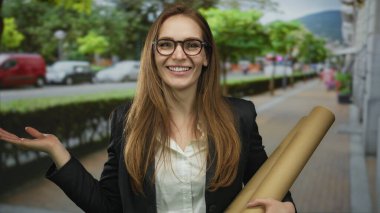 Gülümseyen bir kadın bir elinde karton tüp tutarken diğer elinde el hareketleriyle sokaklara el sallıyor..