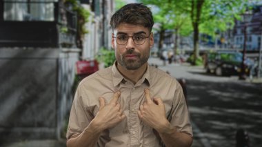 Gündüz vakti şehir merkezinde, gözlüklü, genç İspanyol bir adam baş parmağıyla göğsüne vuruyor..