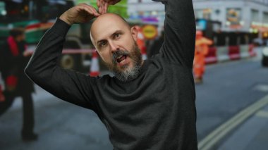 Sakallı kel adam, şehir sokağında güvenle poz veriyor. Araçların geçmişi bulanık ve şehir merkezinde insanlar var..