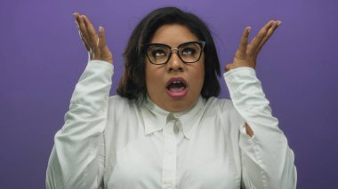 Gözlüklü kadın, mor arka planda ellerini kaldırmış, şok halinde. Beyaz bir gömlek giyiyor. Duygularını ve şaşkınlığını canlı bir arkaplanda aktarıyor..