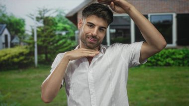 İspanyol asıllı genç adam bina girişinde elleri ile çerçeve hareketi yapıyor; güven ve neşe.