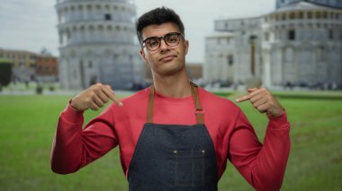 Genç adam gözlüklü ve önlüklü, açık hava sidikli sembolik kulesinin önünde jest yapıyor..
