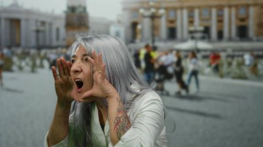 Gri saçlı kadın Vatikan 'ın St. Peter Meydanı' nda duruyor, beyaz bir gömlek giyiyor, açık havada duygularını simgesel İtalyan mimarisi arka planıyla ifade ediyor..