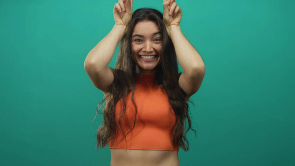 Smiling hispanic woman wearing orange top making bunny ears gesture with fingers in studio; playfulness.
