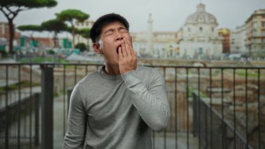 Antik Roma harabelerinin önünde esneyen genç Çinli bir adam, arka planda tarihi mimari olan bir şehir ortamında can sıkıntısını dile getiriyor..