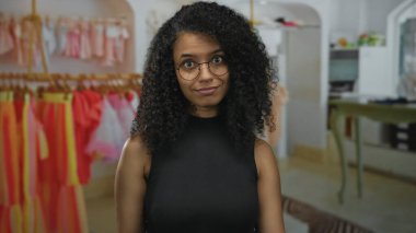 Bir giyim mağazasının içinde renkli raflar arasında parmaklarını dudaklara doğru hareket ettiren genç Latin kadın; gizli sessizlik.