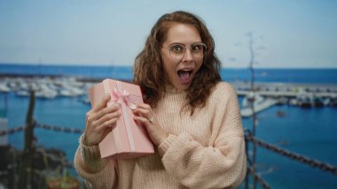Sahilde pembe hediye kutusuyla gülümseyen, gözlüklü, süveterli, dışarıda duran, arka planda bulanık bir limanı olan bir kadın, mutluluğu işaret ediyor..