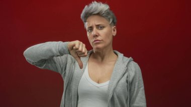 Kırmızı stüdyoda gri kapüşonlu kısa gri saçlı bir kadın baş parmak hareketi gösteriyor; reddedilme hüsranı.