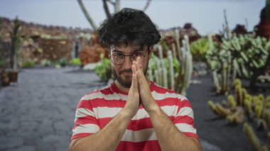 Sakallı genç adam açık hava kaktüs bahçesi parkında dua ediyor. Çizgili gömlek ve gözlük takıyor. Açık mavi gökyüzünün altında odaklanmış ve sakin görünüyor..