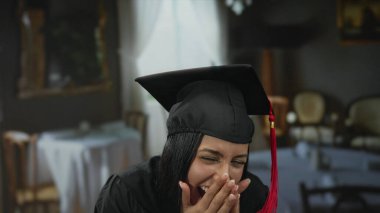 Mezuniyet şapkalı ve cüppeli genç İspanyol kadın ışıkla dolu bir restoran ortamında gülüyor ve puan veriyor, neşeli bir anı kutluyor..