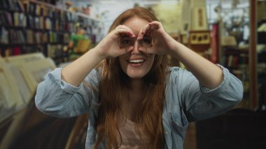 Parlak gülüşlü genç beyaz kadın kütüphanede el dürbünü hareketi yapıyor..