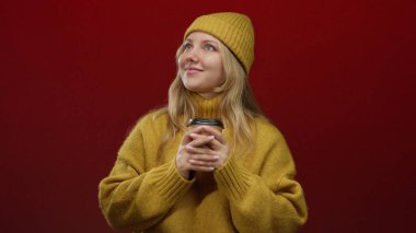 Sarı süveter ve şapka giyen genç sarışın bir kadın elinde kahve fincanıyla kırmızı bir fonda gülümsüyor..