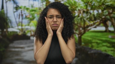 Genç İspanyol kadın, güneş ışığında yeşil bir parkta kıvırcık saçlarına dokunurken birbirine dolanmış tellere kaşlarını çatıyor..