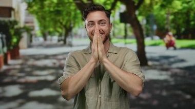 Sakallı genç bir adam yazın güneşli bir sokakta arka planda yeşil ağaçlarla gülümsüyor, rahat bir gömlek giyiyor ve neşeli bir ifade sergiliyor.