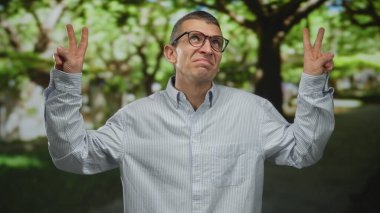 Parkta gözlüklü bir adam hava tırnak içinde iğneleyici sözler sarf ediyor ve açık havada yemyeşil bir ortamda günlük kıyafetler giyiyor..