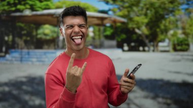 Kırmızı kazaklı genç adam, açık hava parkında ağaçlarla çevrili bir yerde akıllı bir telefon tutarak gülümsüyor. Neşeli bir doğal ortamda güneşli bir günde rahatlatıcı ve mutlu bir an yaşıyor..