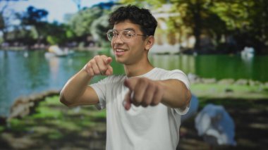 Göl kenarındaki yeşil orman parkında gözlüklü bir adam kamerayı işaret ediyor..