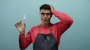 Gözlük ve önlük takan genç adam, kuaför ya da kuaför olarak yeteneklerini gösteren izole edilmiş mavi duvar arkaplanına karşı makas ve makas tutuyor.
