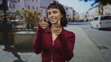 Gülümseyen yüzlü genç İspanyol kadın şehir caddesinde kamerayı işaret ediyor..