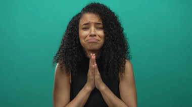 Kıvırcık saçlı ve yalvaran ifadeli bir kadın canlı yeşil bir arka plana karşı tek başına duruyor, dramatik bir duygu ve umut sahnesi yaratıyor..