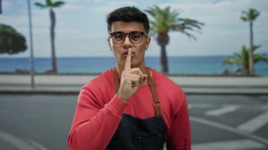 Gözlüklü ve önlüklü genç adam deniz kenarında palmiye ağaçları ve okyanus manzaralı bir yerde sessizlik jesti yapıyor.