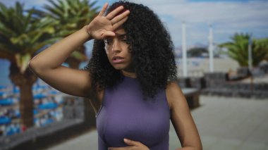 Sahilde rahatsız olduğunu ifade eden bir kadın. Açık havada, güneşin altında palmiye ağaçları var..