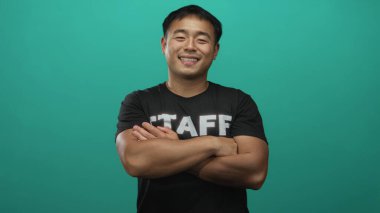 Çapraz kollu siyah bir personel tişörtü giyen bir adam ve bir gülümseme ve bir gök mavisi stüdyosunda doğrudan bakış; güven.