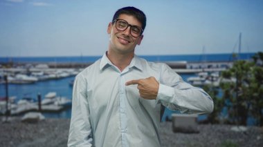 Gözlüklü bir adam iskelede teknelerin yanında dikiliyor. Parmakla göstererek gömleği işaret ediyor. Kendine güveni tam..