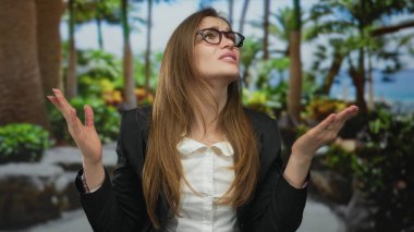 Gözlük takan ve spor ceket giyen genç bir kadın ormanda yeşil yaprakların arasında yukarı bakıyor..