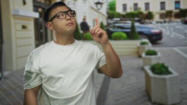 Genç Çinli gözlüklü ve beyaz tişörtlü bir adam, parmakla parmakla gösterilen kaldırımda yukarı doğru, park halindeki arabanın yanında, merak.