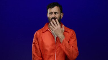 Turuncu hapishane tulumu giyen İspanyol sakallı adam, mavi bir arkaplana dokunduğu için endişeli olduğunu ifade ediyor. Tutukluluk ve endişe temalarını çağrıştırıyor..