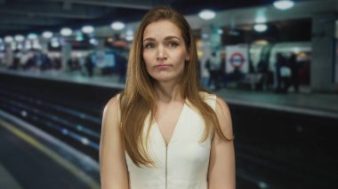 İstasyondaki tren raylarının yanında çıplak kollarıyla ayakta duran kadın tarafsız bir ifadeyle, sabırsız..